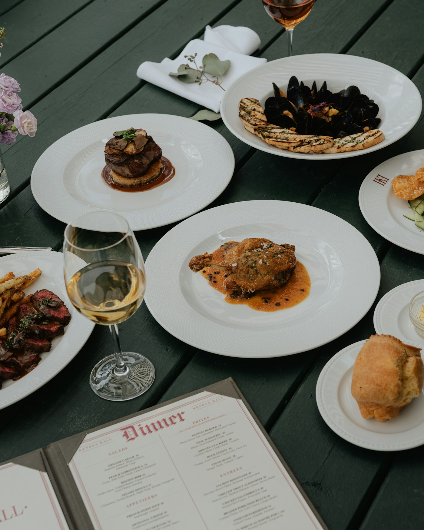 Plated dinner dishes on a dark wooden table with wine glasses and an open menu